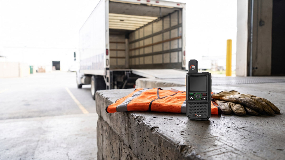 An Echo radio stands on the edge of a loading dock beside a folded high-visibility vest, with a delivery truck backed up to the bay behind it.