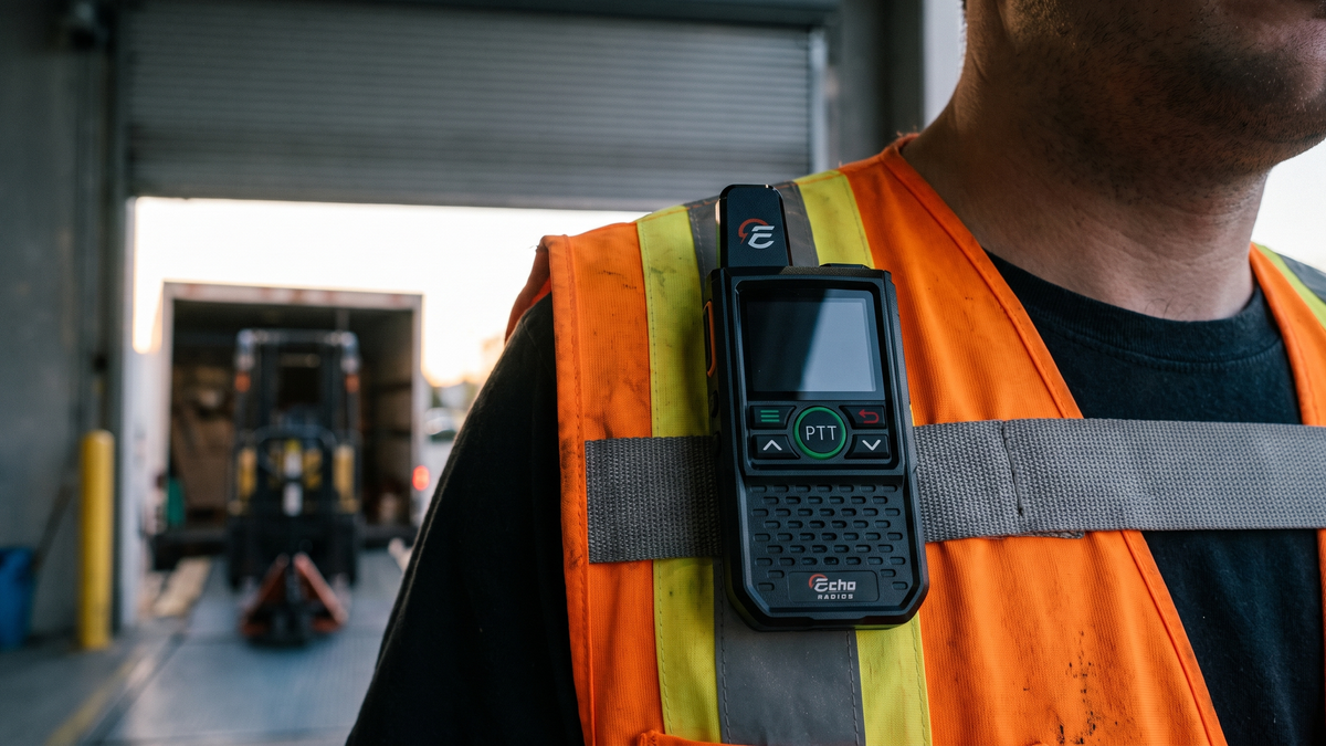 A compact Echo radio clipped to the chest strap of a plain work vest, with a loading bay blurred behind at dawn.