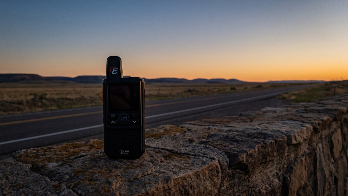 A compact handheld radio stands on a stone wall at a highway overlook at dusk while an empty road runs straight to the horizon behind it.