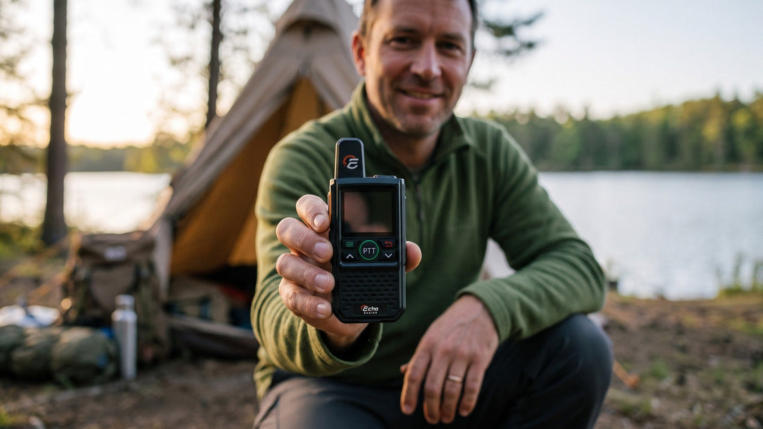 At a lakeside campsite, a man holds a compact handheld radio out toward the camera, its green push-to-talk button facing forward.