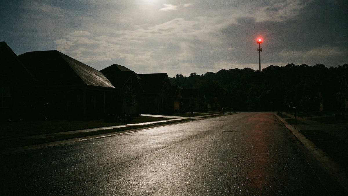 A dark suburban street at night in a power cut, with one red light glowing on a cell tower on the ridge behind the houses.