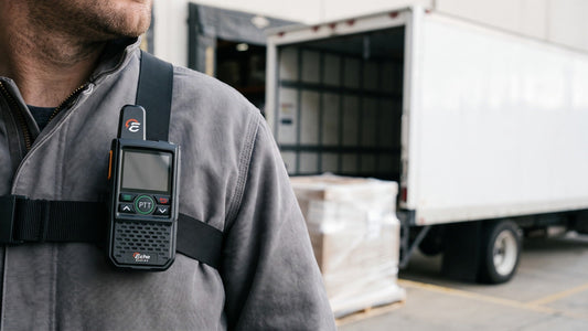 A compact handheld radio clipped to the chest strap of a work jacket at a warehouse loading dock, a box truck's open side door behind.